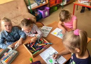 Świetliczaki na zajęciach z języka niemieckiego - „Każdy dobrze o tym wie, język niemiecki przyda się!”. Na stoliku leży pudełko z kolorowymi pisakami. Dzieci siedzą przy stole i kolorują wydrukowane wzory.
